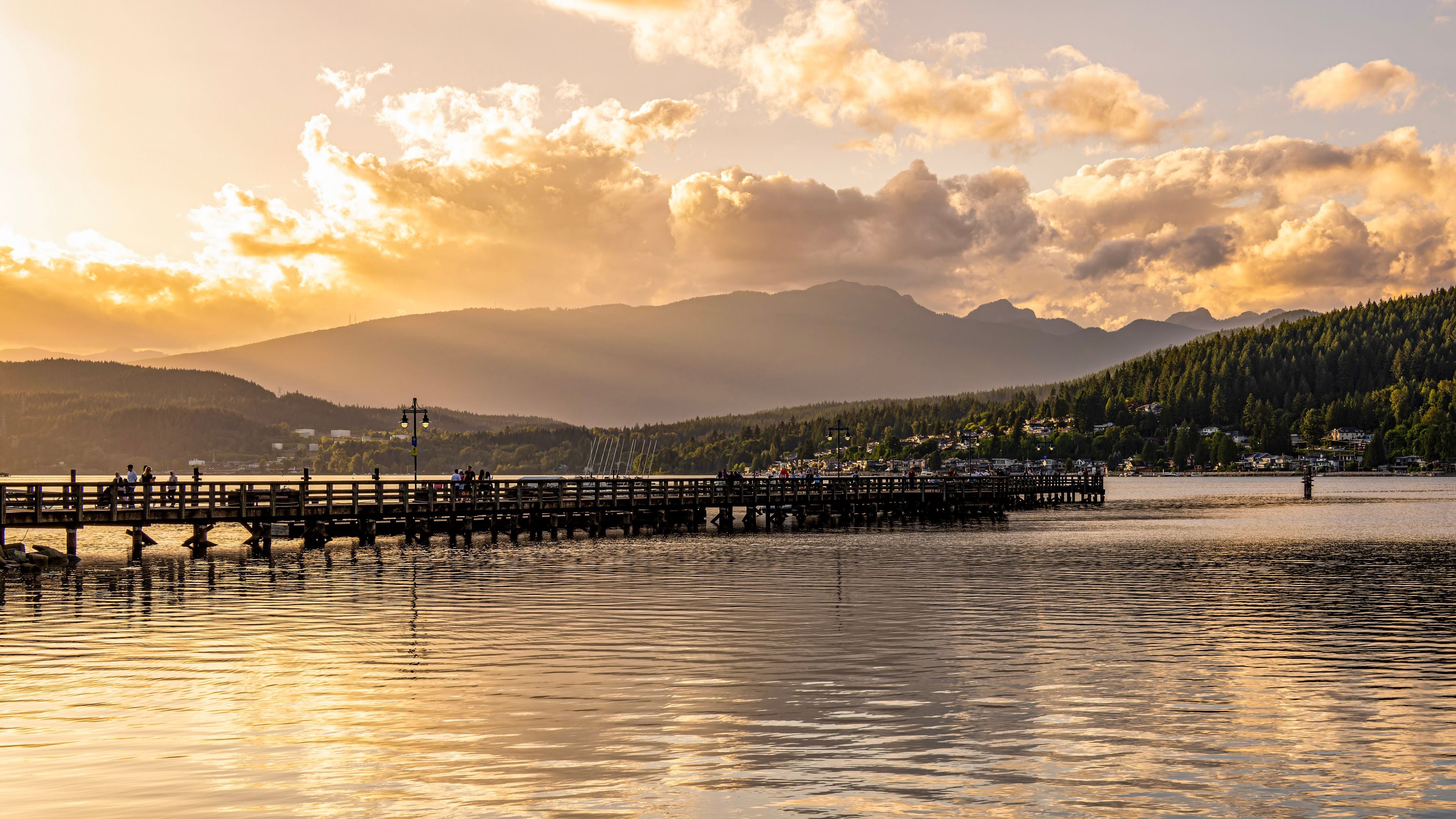 A sunset over a lake with a wooden pier extending into the water, surrounded by mountains and a forested hillside.
