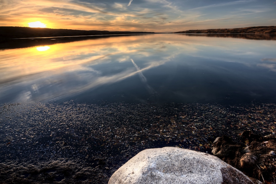 Un lago tranquilo al atardecer con un reflejo claro del cielo, rodeado de terreno rocoso.