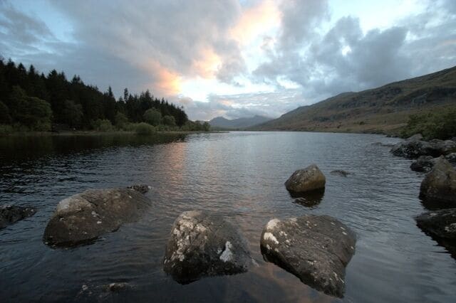 A calm river with large rocks in the foreground, surrounded by hills and a forested area.