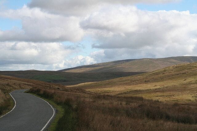 A winding road through a hilly landscape with grassy fields and a cloudy sky.