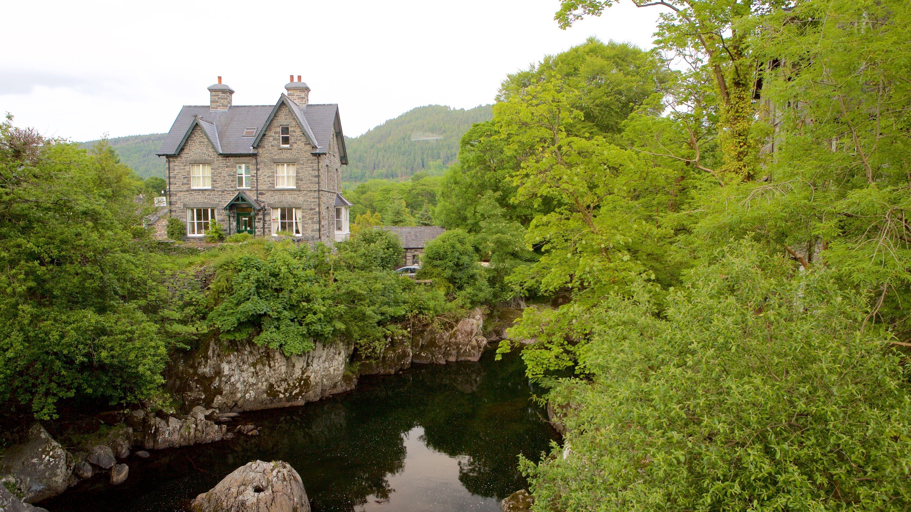 A stone house with a steep roof, surrounded by lush greenery and a river.