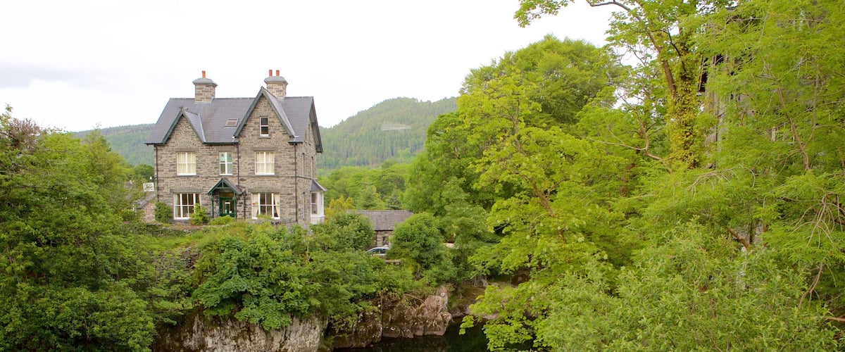 A stone house with a steep roof, surrounded by lush greenery and a river.