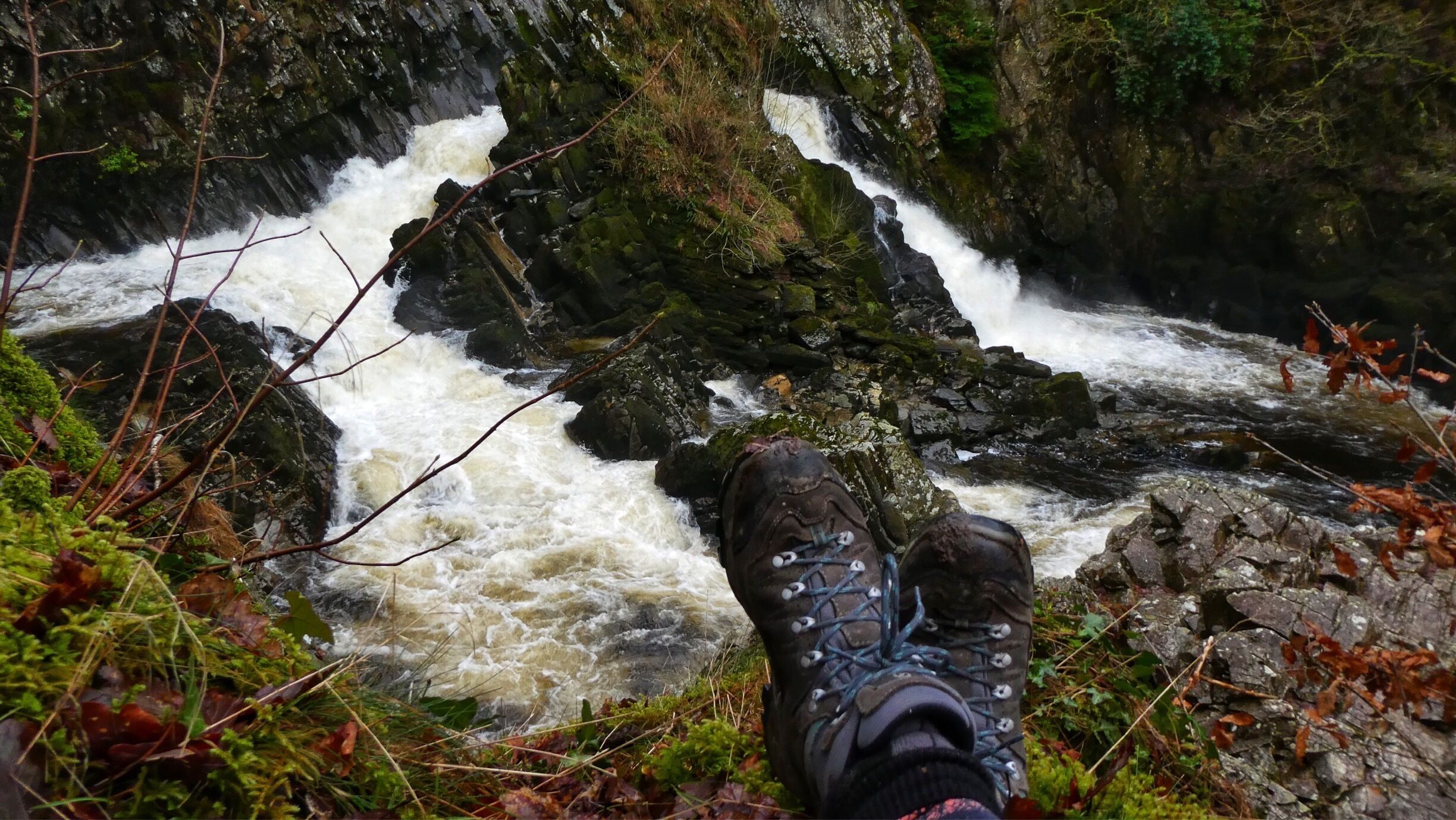 A person's foot in a hiking boot overlooking a fast-flowing river with rocky terrain and greenery.