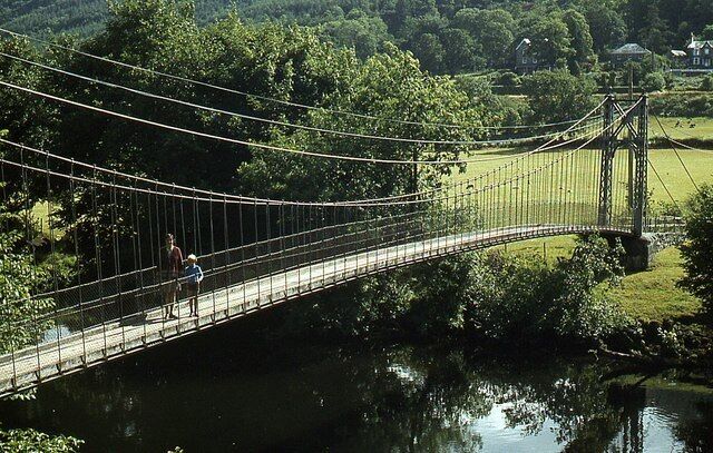 Un puente colgante sobre un río, rodeado de vegetación exuberante y un pequeño edificio al fondo.