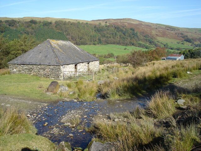 A stone house with a thatched roof in a rural landscape with a stream and rolling hills in the background.