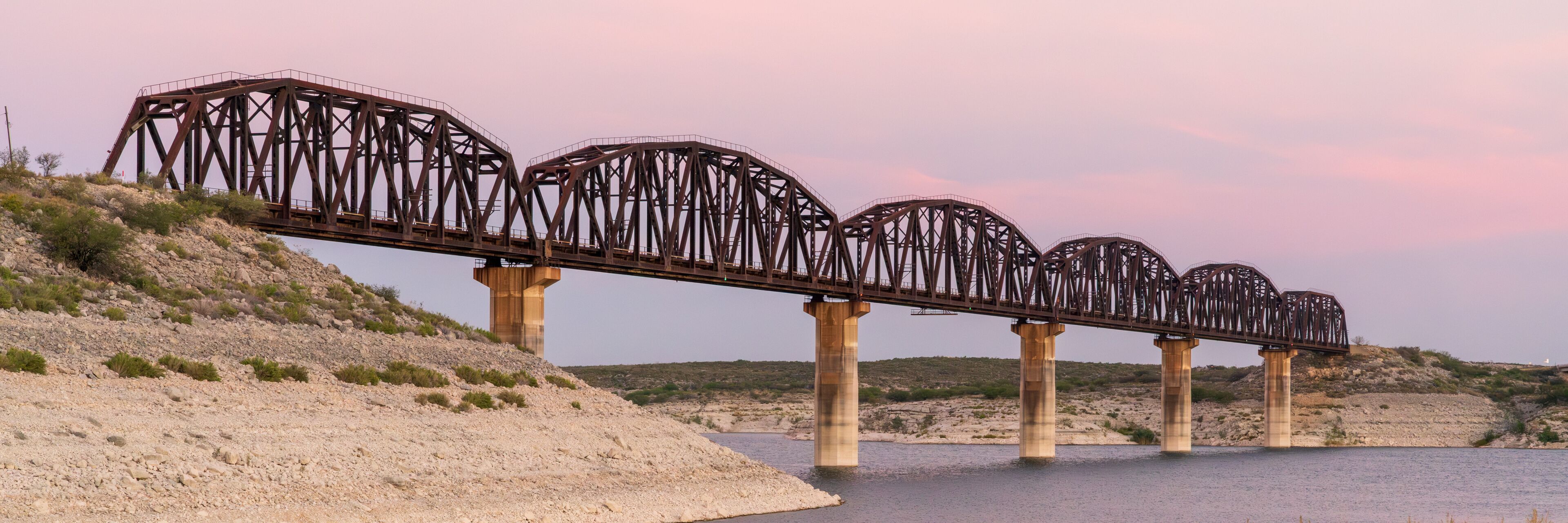 A steel bridge with multiple arches spanning a river, supported by concrete pillars, set against a hilly landscape with sparse vegetation.