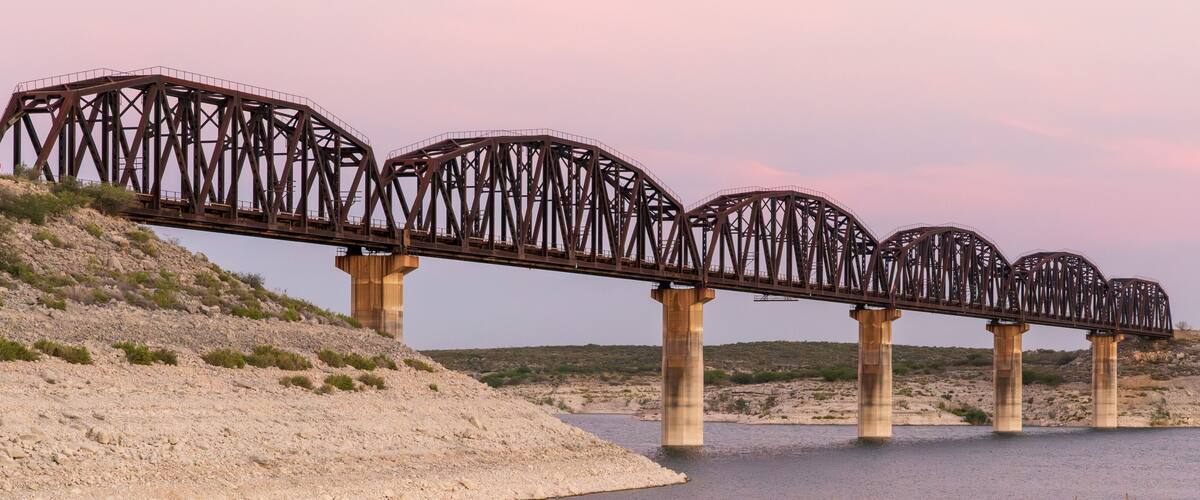 A steel bridge with multiple arches spanning a river, supported by concrete pillars, set against a hilly landscape with sparse vegetation.