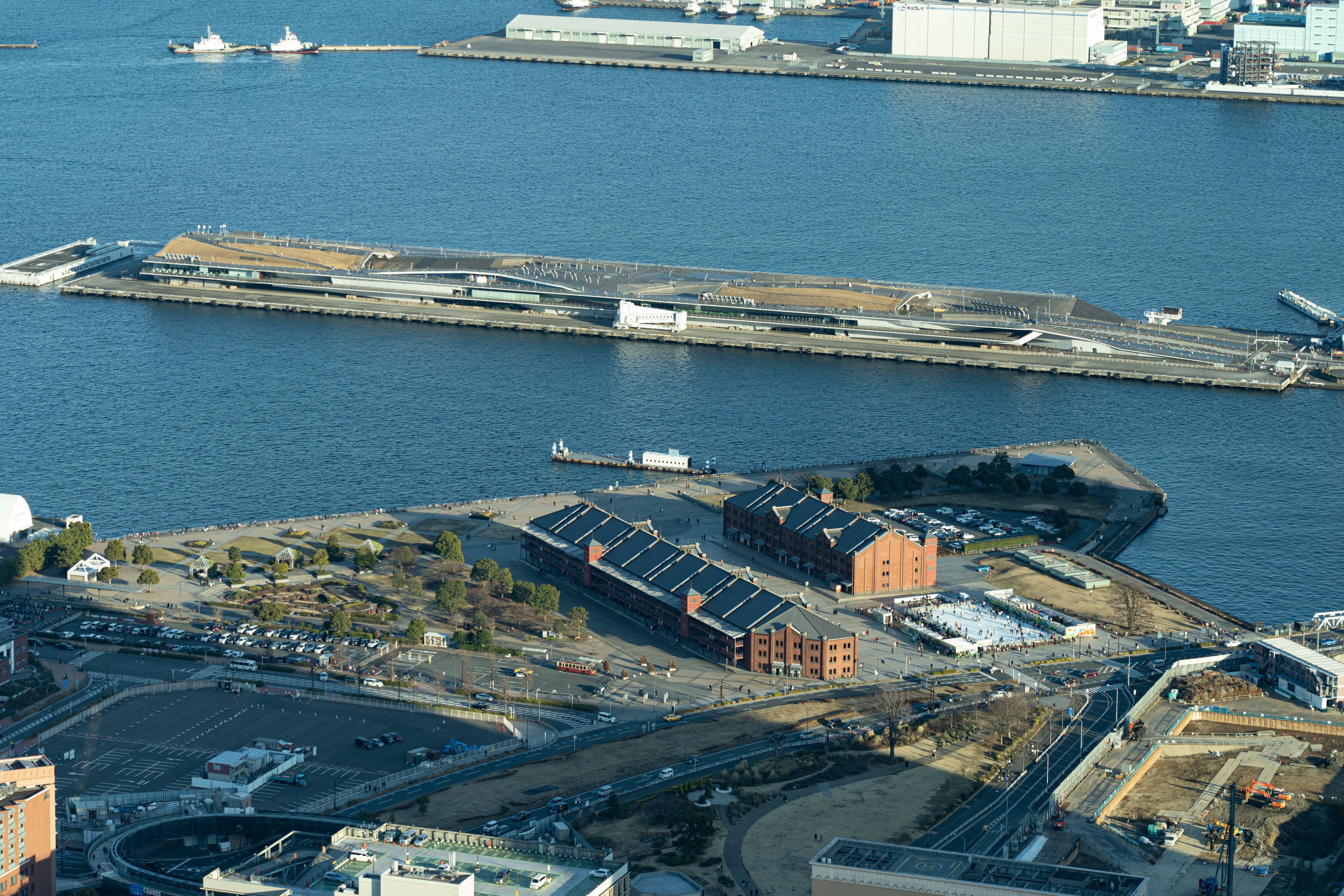 An aerial view of a port area with ships, a large industrial building, and a bridge.