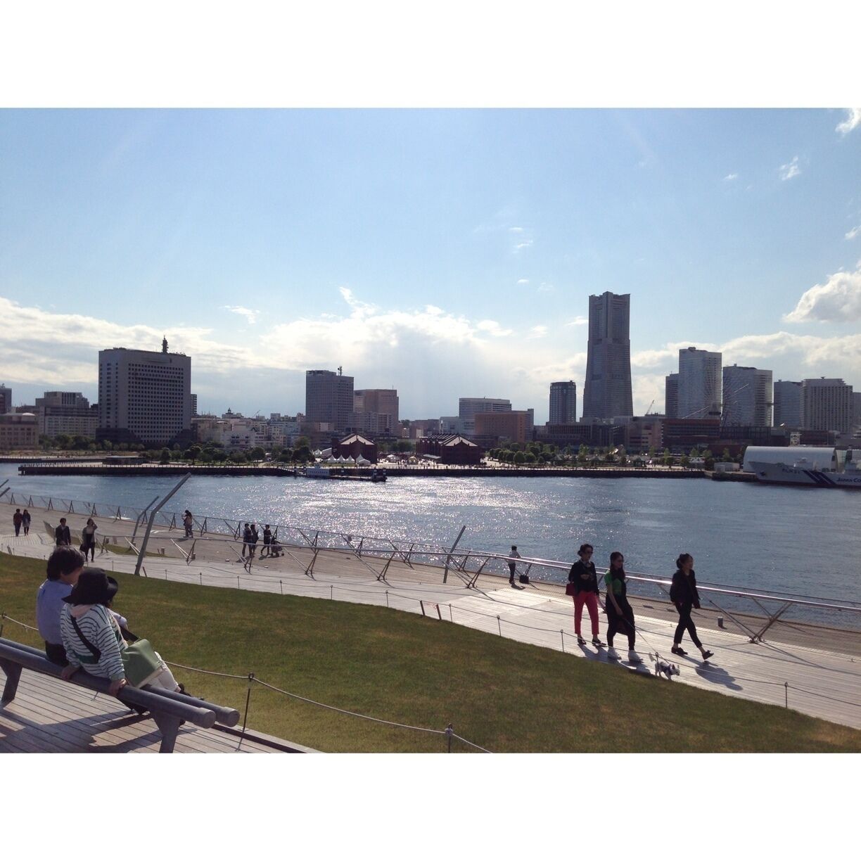Un paseo fluvial con gente caminando y sentada, un horizonte urbano al fondo y un cielo despejado.