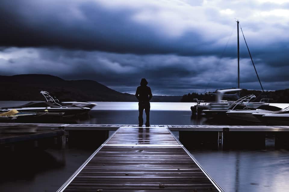 A person standing on a wooden dock facing a calm lake with boats docked on the right and mountains in the background.