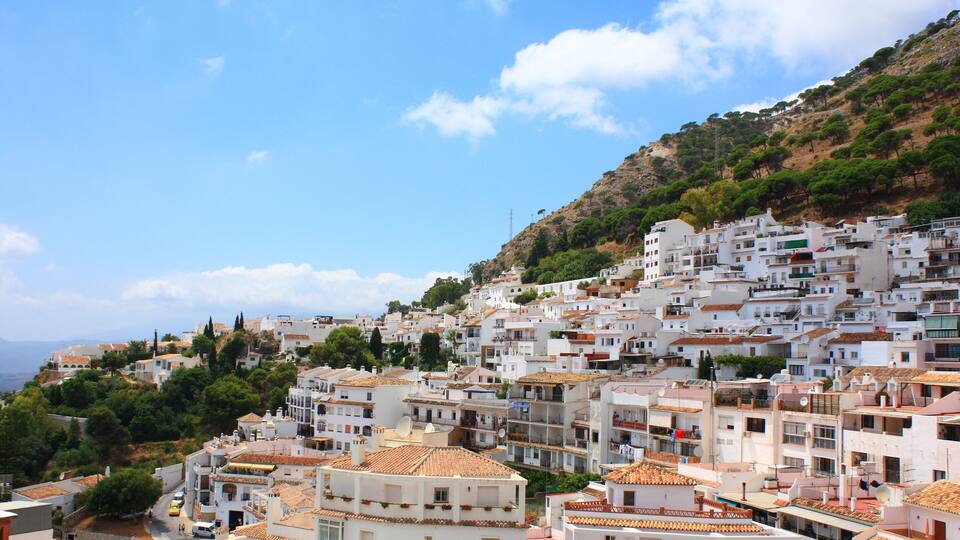 A densely packed hillside town with white buildings and terracotta roofs, surrounded by greenery and a clear blue sky.