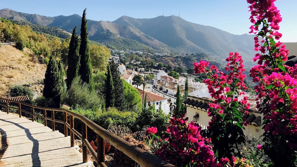 Ein malerischer Blick auf eine Stadt, eingebettet in ein Tal, mit Bergen im Hintergrund, einer hölzernen Treppe und leuchtend rosa Bougainvillea-Blüten.