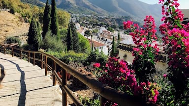 Vista pitoresca de uma vila aninhada num vale, com montanhas ao fundo, uma escadaria de madeira e flores vibrantes de buganvília rosa.