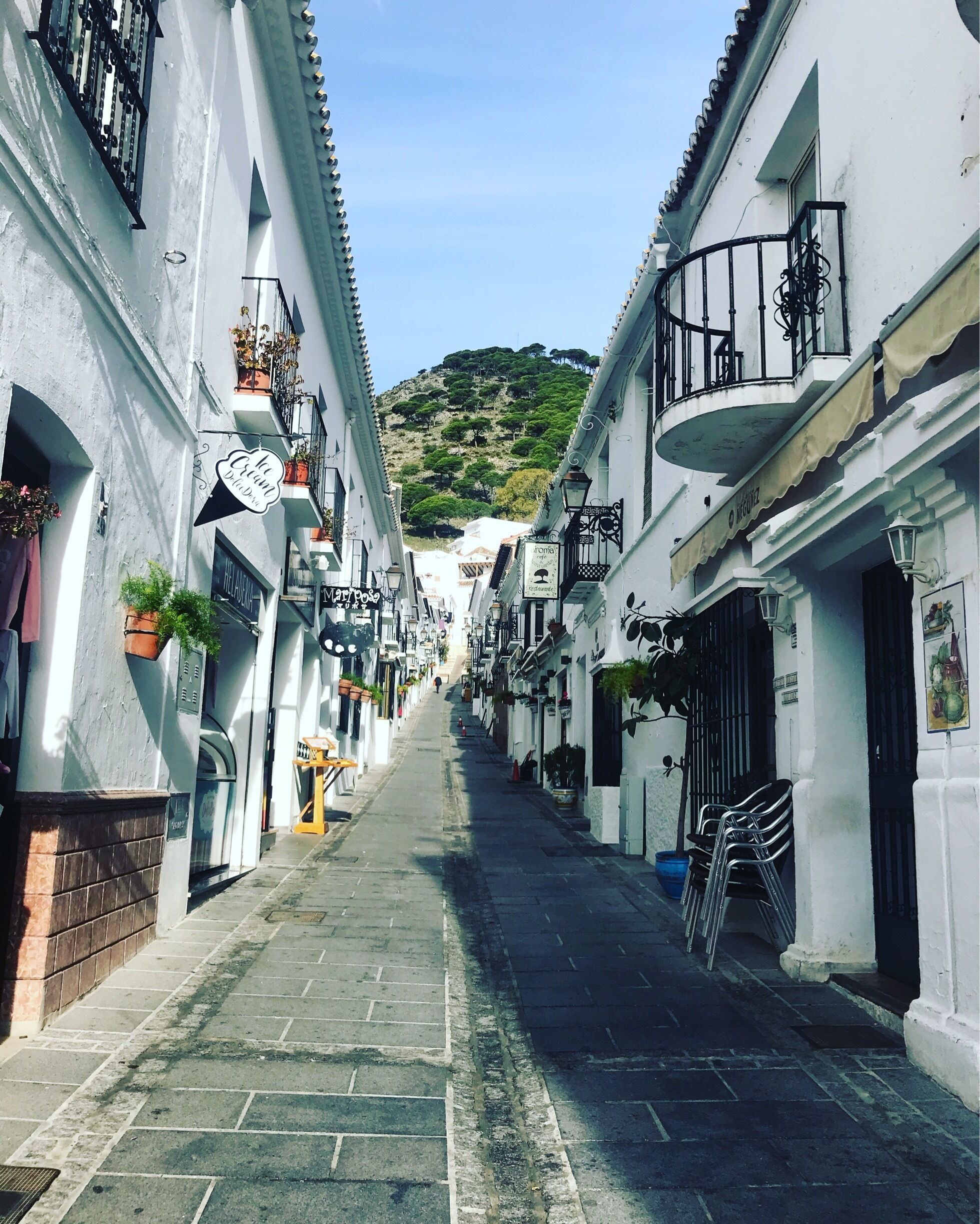 A narrow street flanked by white buildings with balconies and hanging plants, leading up a hill.