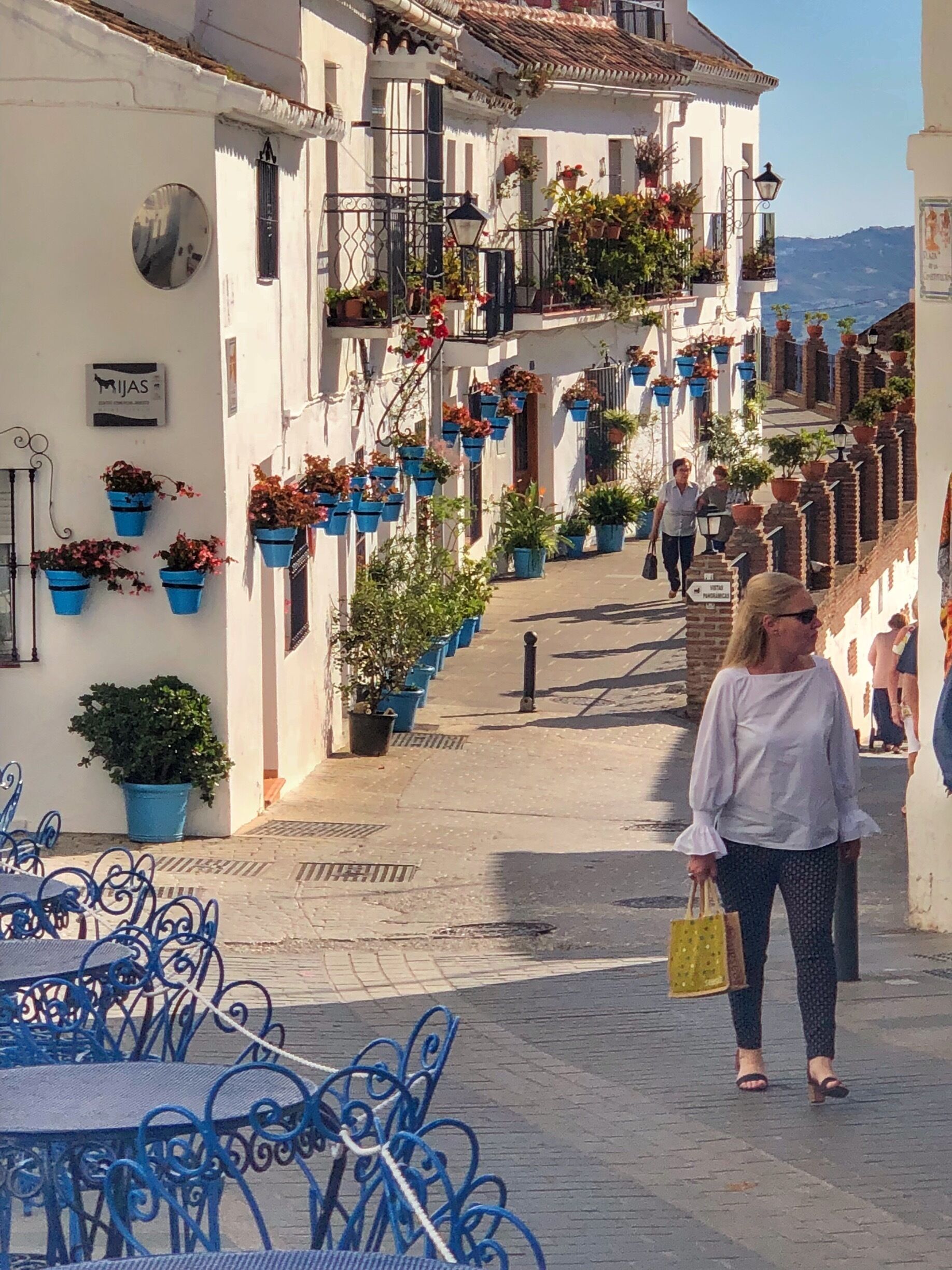 A narrow street lined with white buildings, blue outdoor seating, and potted plants.
