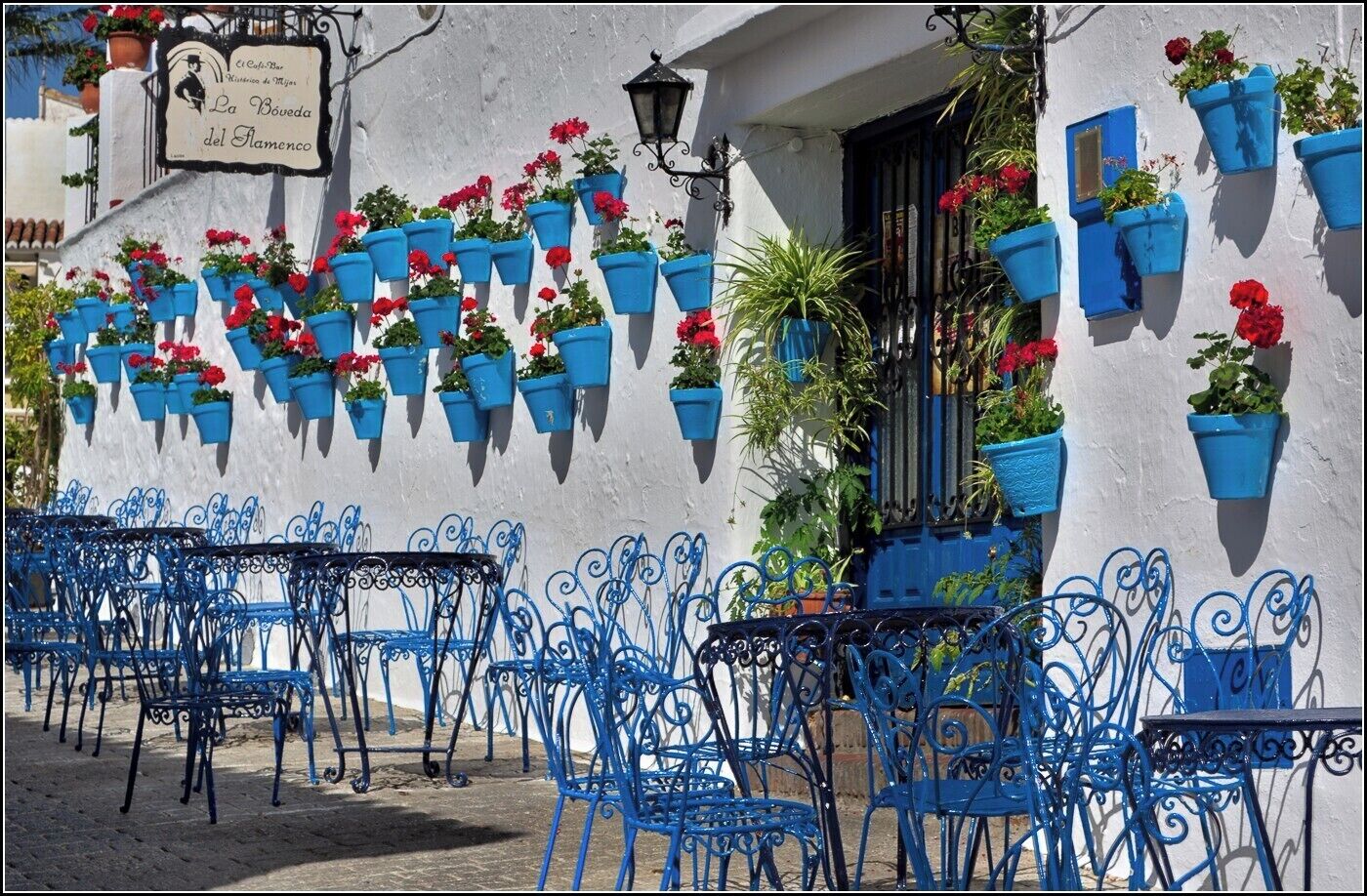 Outdoor seating with blue metal chairs and tables, blue hanging flower pots, and a building with a sign.