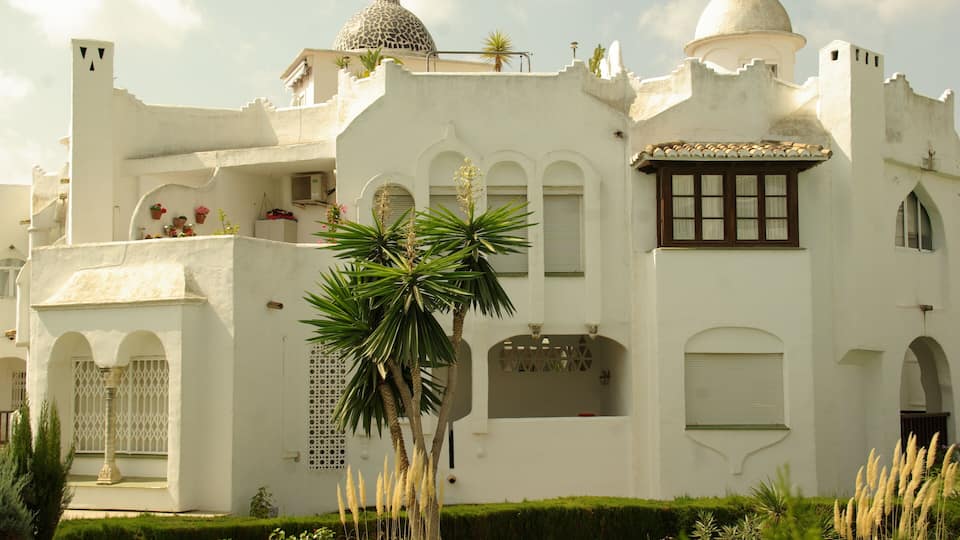 A white building with domes, arched windows, and a garden with palm trees.