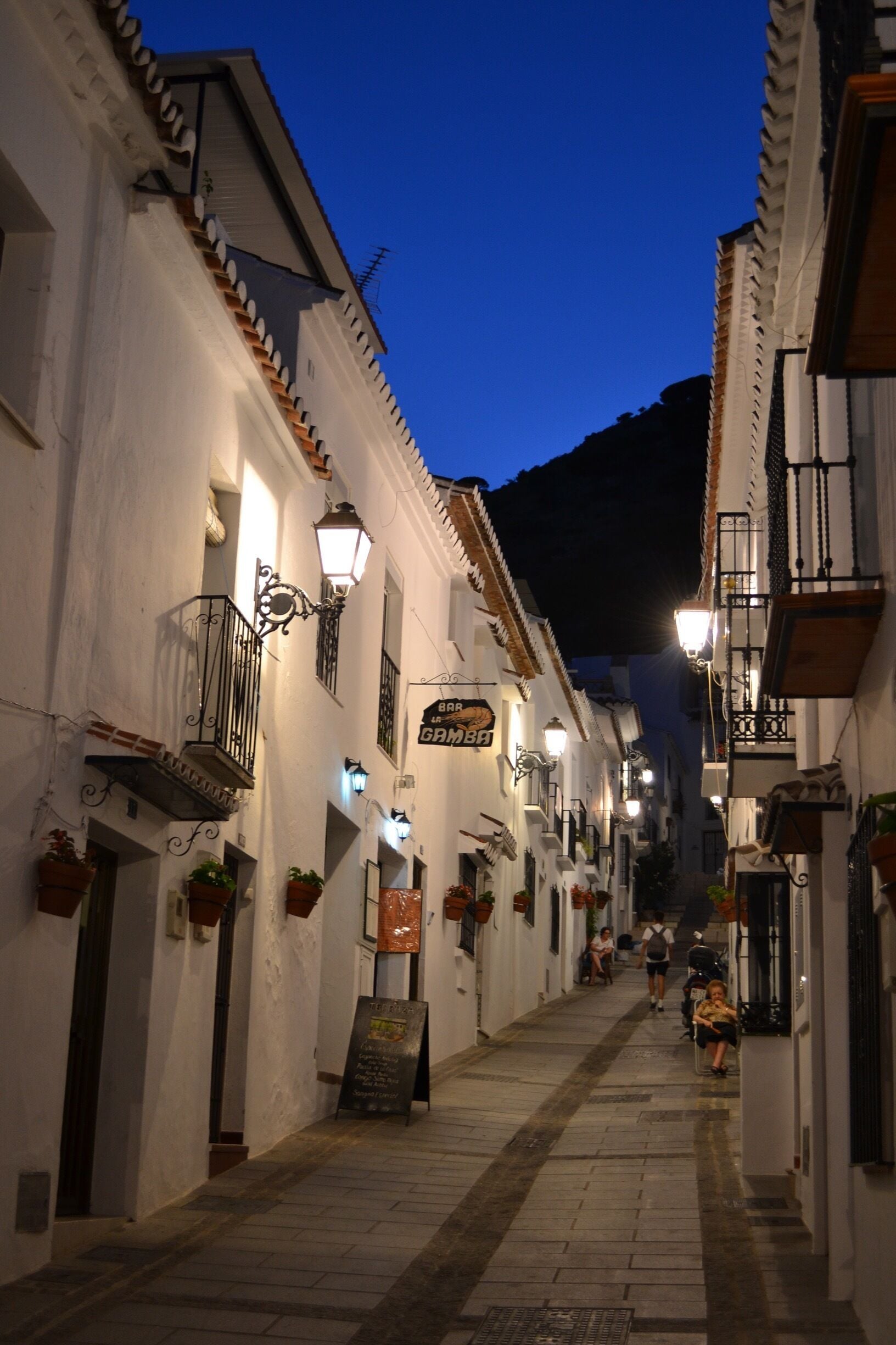 A narrow street with white buildings, outdoor seating, and hanging plants.