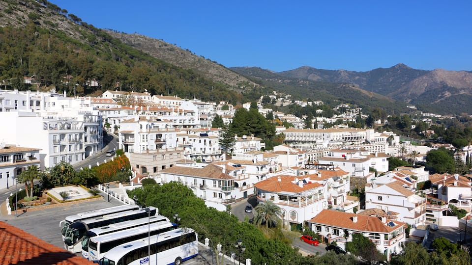 A coastal town with white buildings, a bus, and a mountainous backdrop.