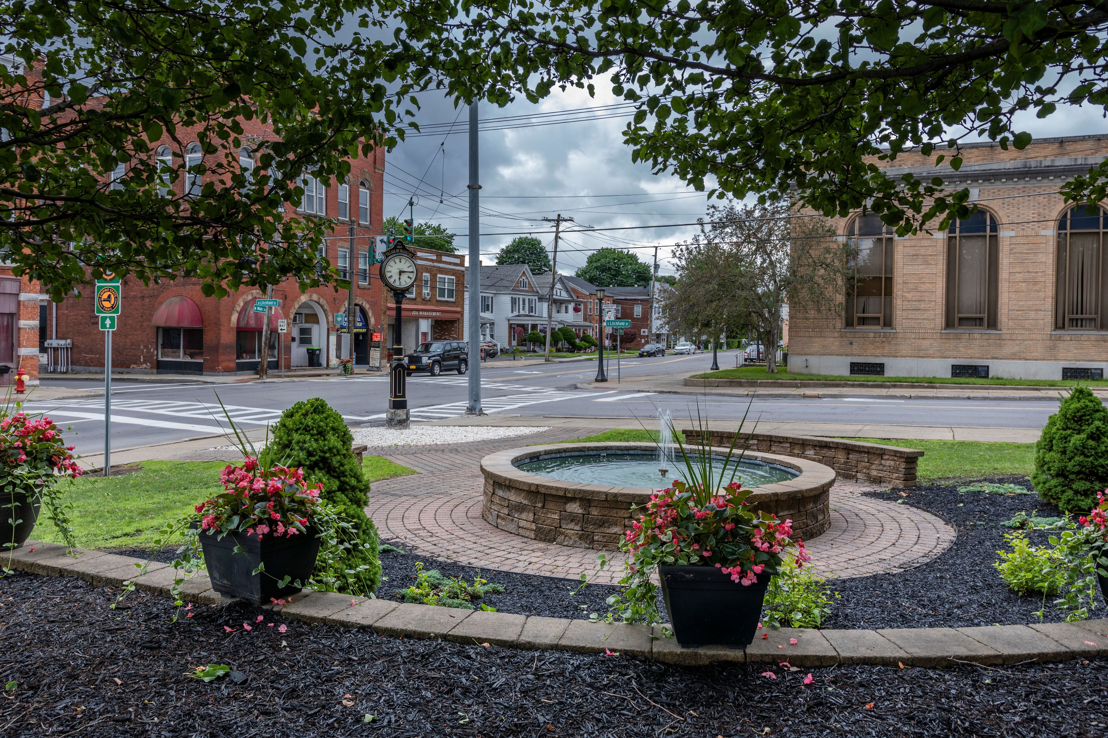 A city intersection with a roundabout, brick buildings, and a fountain surrounded by flower beds.