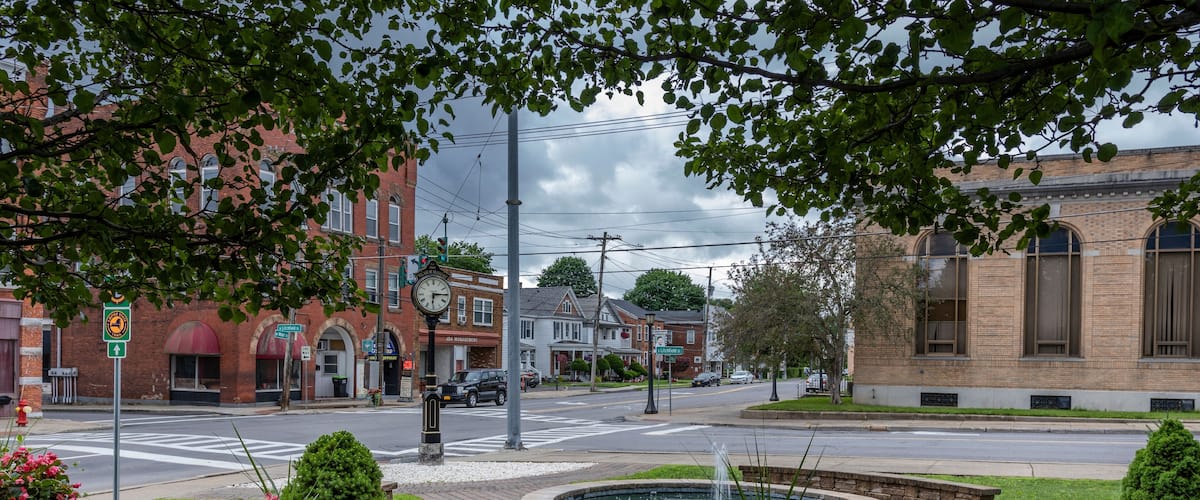 A city intersection with a roundabout, brick buildings, and a fountain surrounded by flower beds.