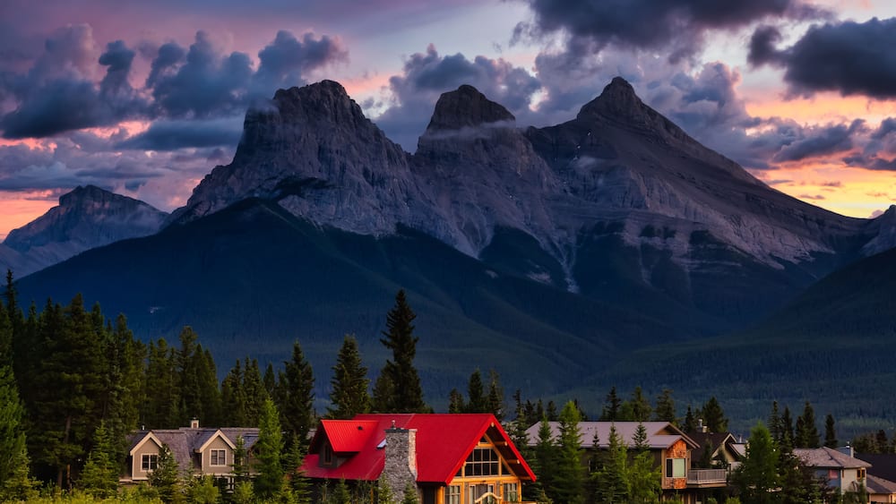 A mountain range with a prominent peak, a red-roofed cabin, and several other houses in a forested valley.
