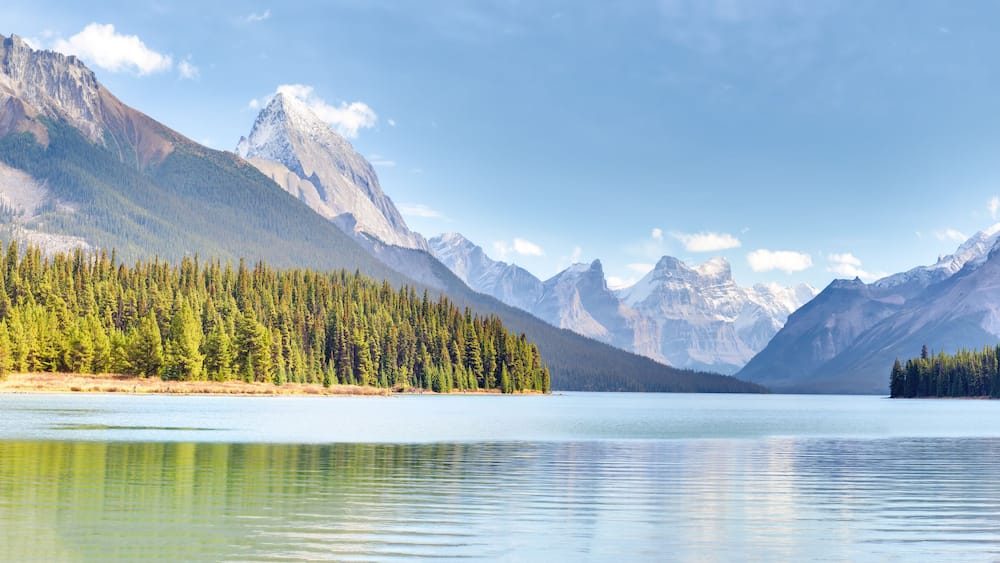 A calm lake surrounded by dense forests and snow-capped mountains under a clear sky.