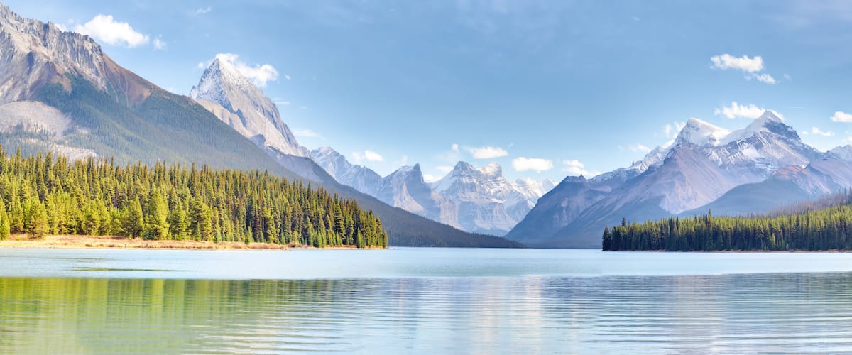 A calm lake surrounded by dense forests and snow-capped mountains under a clear sky.