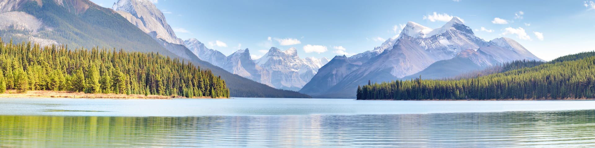 A calm lake surrounded by dense forests and snow-capped mountains under a clear sky.