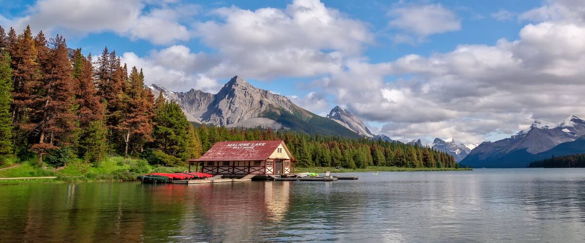 A lakeside cabin with a red roof, surrounded by dense forests and towering mountains.