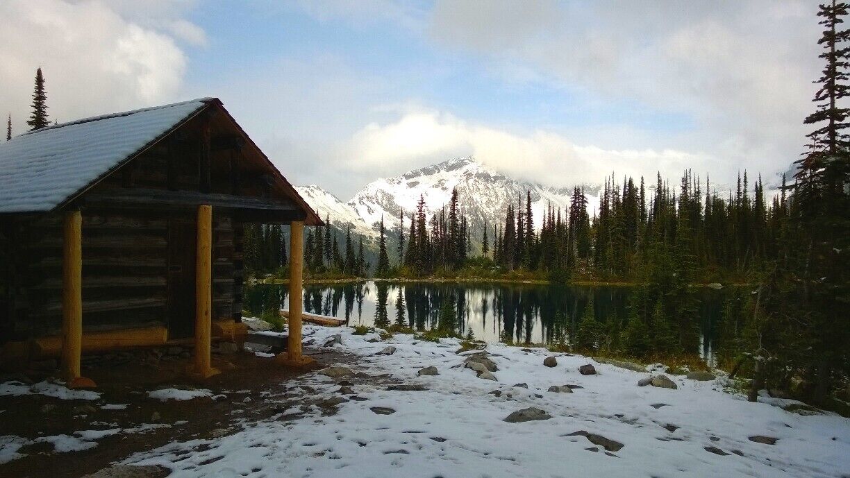 A wooden cabin by a lake with snow-covered ground and a mountain in the background.