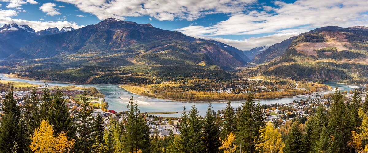 Un paysage montagneux avec une rivière, une ville et des arbres aux couleurs automnales.