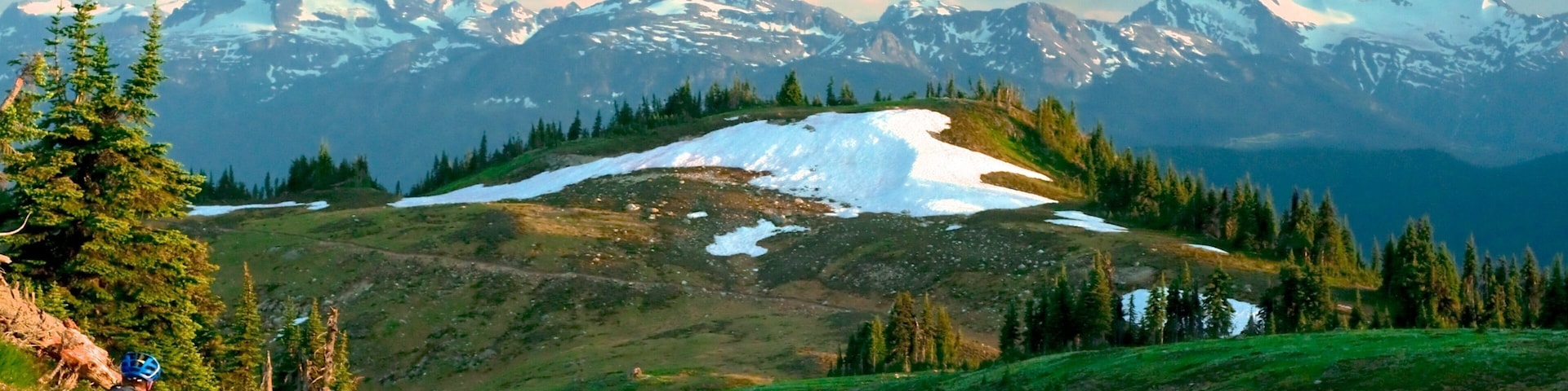 A mountain landscape with a cyclist on a trail, snow patches, and a forested area.