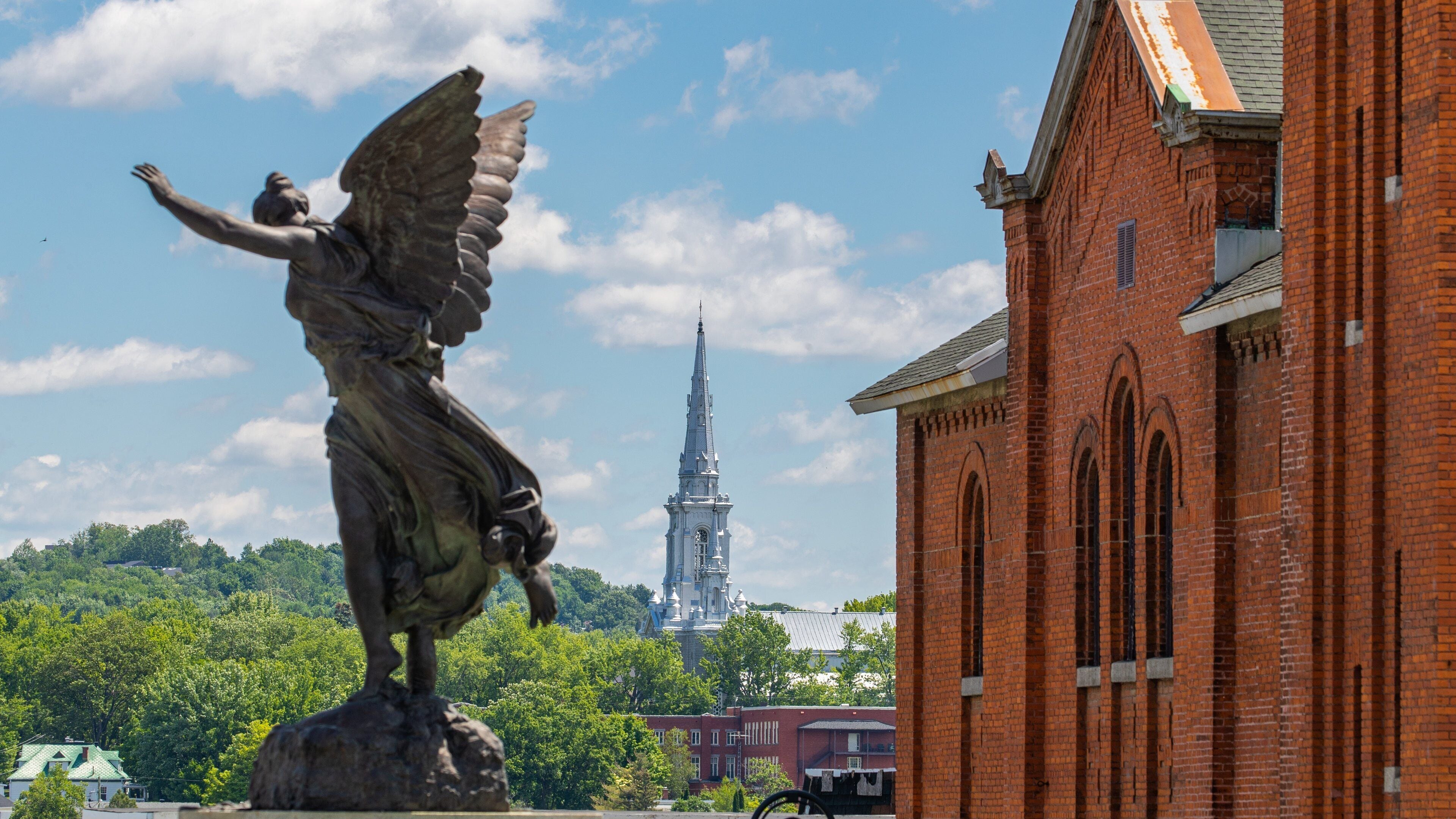 A statue of an angel with outstretched wings, a church with a spire, and a brick building in the foreground.