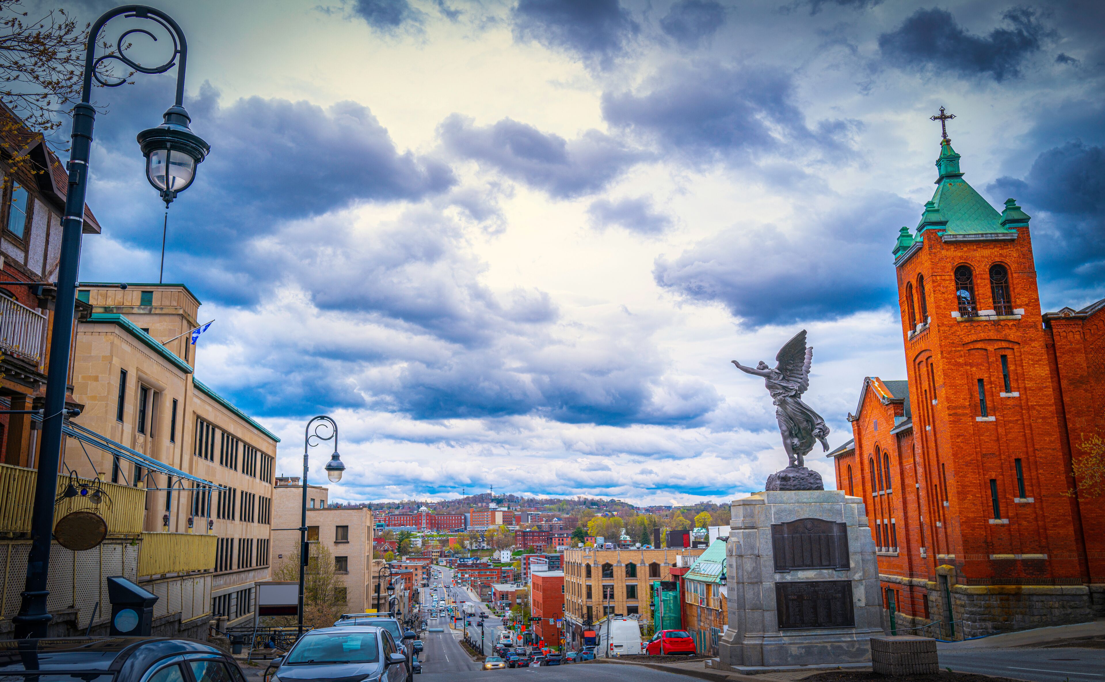 A city street with a statue of an angel, a red brick church, and a cloudy sky.