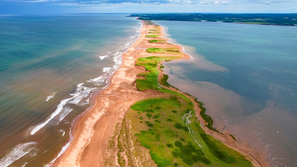 A coastal landscape with a sandy beach, green vegetation, and a clear view of the ocean.