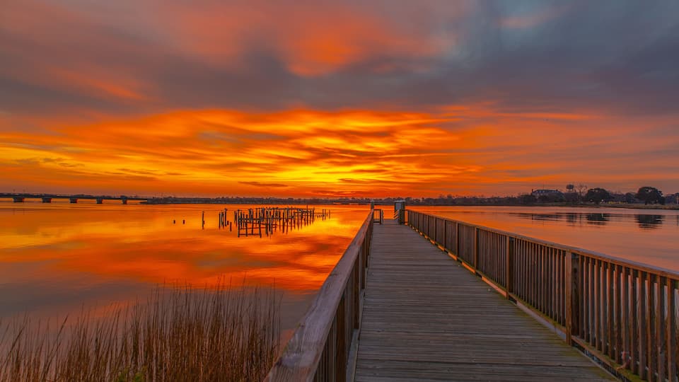 A wooden boardwalk extending over a calm body of water, reflecting a vibrant sunset sky.