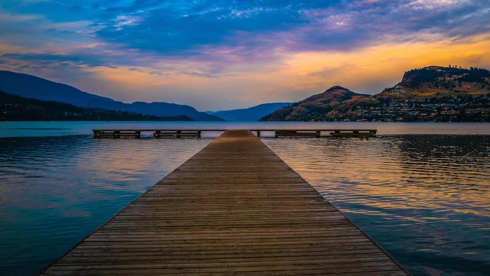 A wooden pier extending into calm waters, surrounded by mountains and a town in the distance.