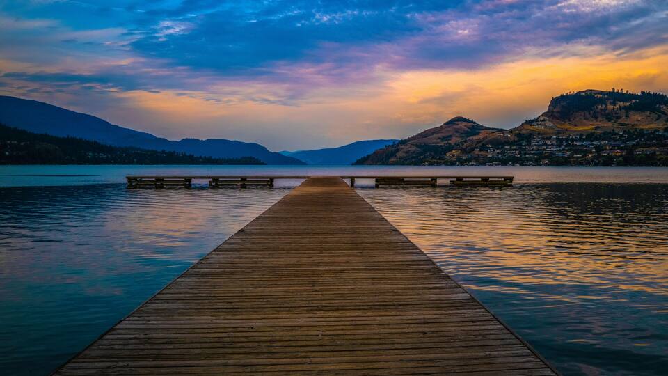 A wooden pier extending into calm waters, surrounded by mountains and a town in the distance.