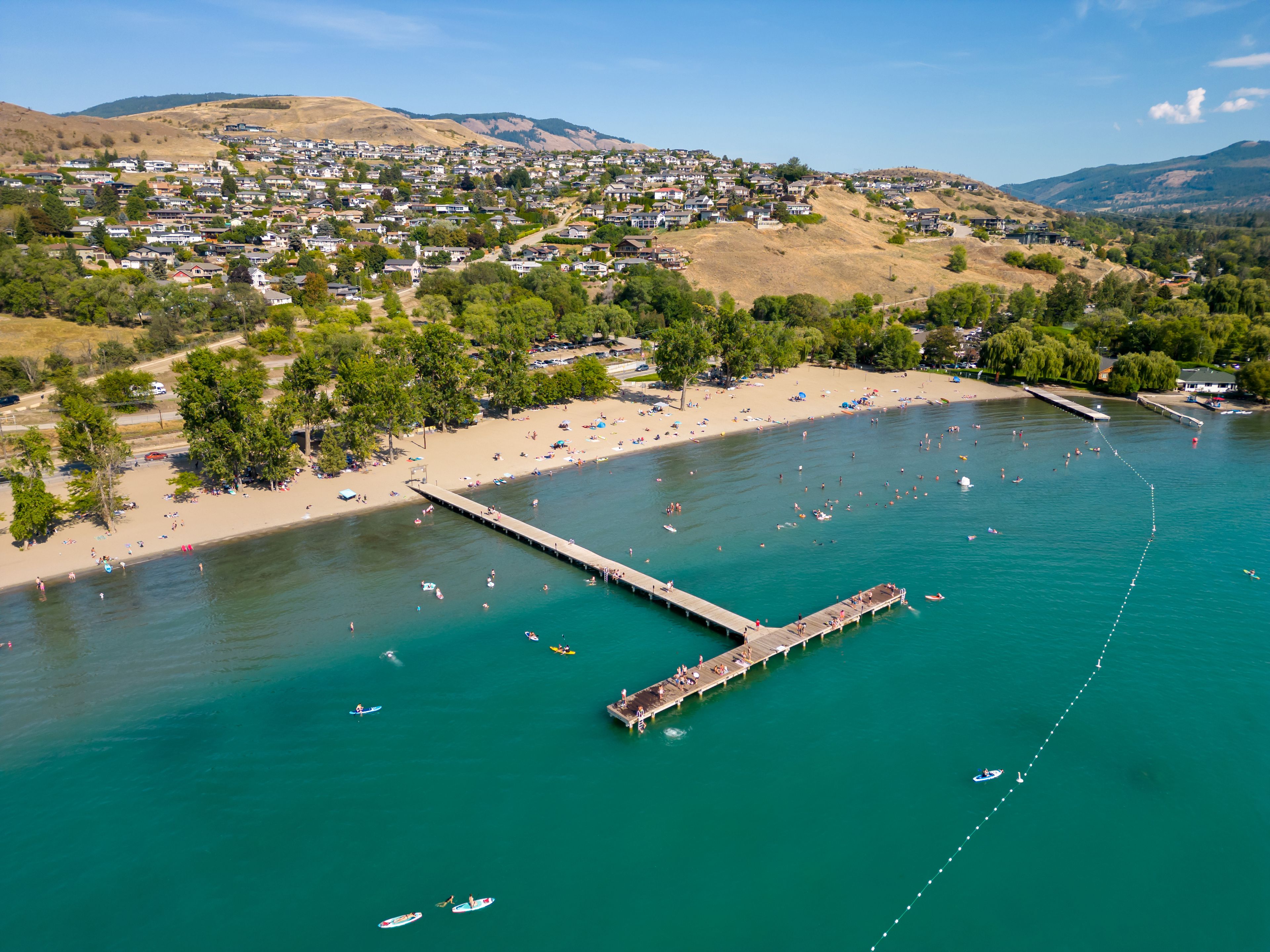 A coastal town with a pier extending into turquoise waters, surrounded by hills and a residential area.