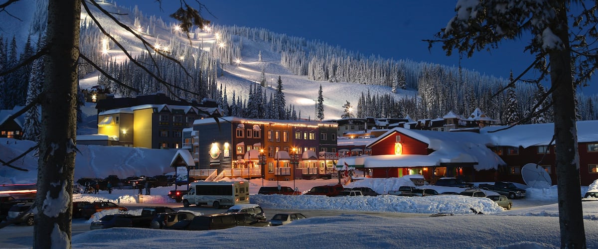 A snow-covered village with illuminated buildings and a ski slope in the background.
