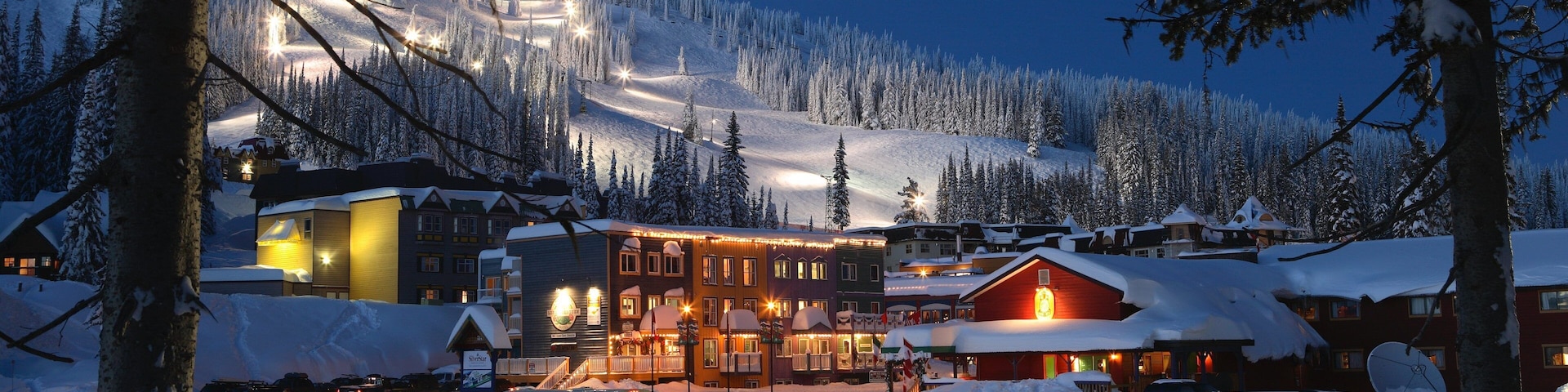 A snow-covered village with illuminated buildings and a ski slope in the background.