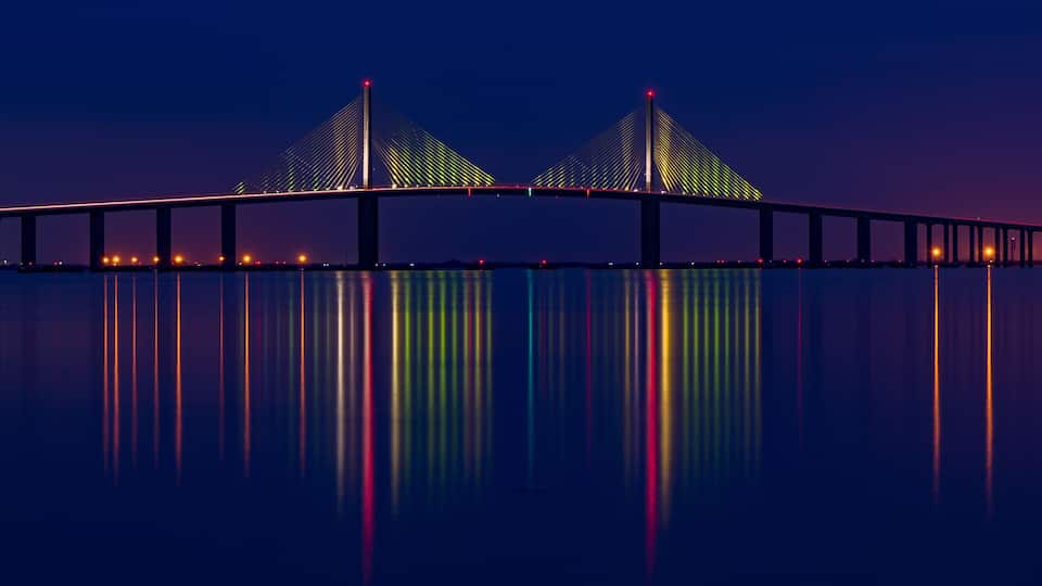 A bridge with two towers and colorful lights reflecting on the water.