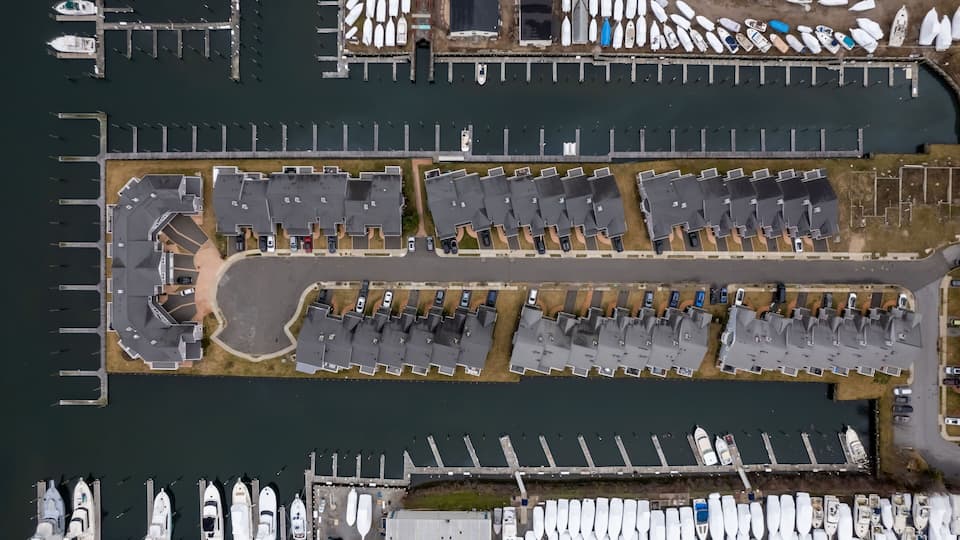 An aerial view of a marina with numerous boats docked, a parking area, and a building with a sloped roof.