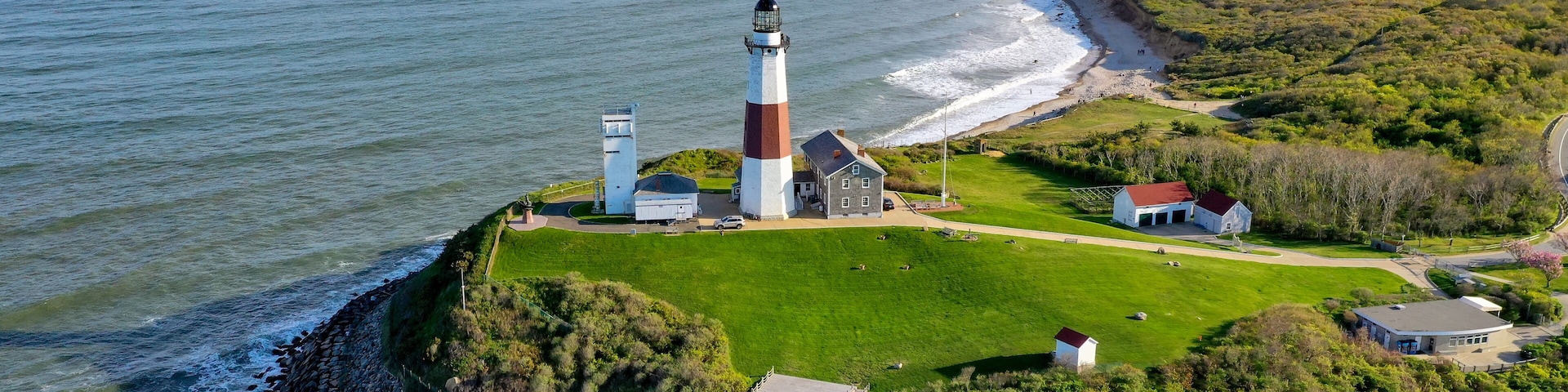 A lighthouse on a rocky outcrop with a grassy area and buildings nearby.