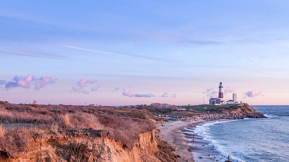 A coastal landscape with a lighthouse, grassy dunes, and a rocky shoreline.
