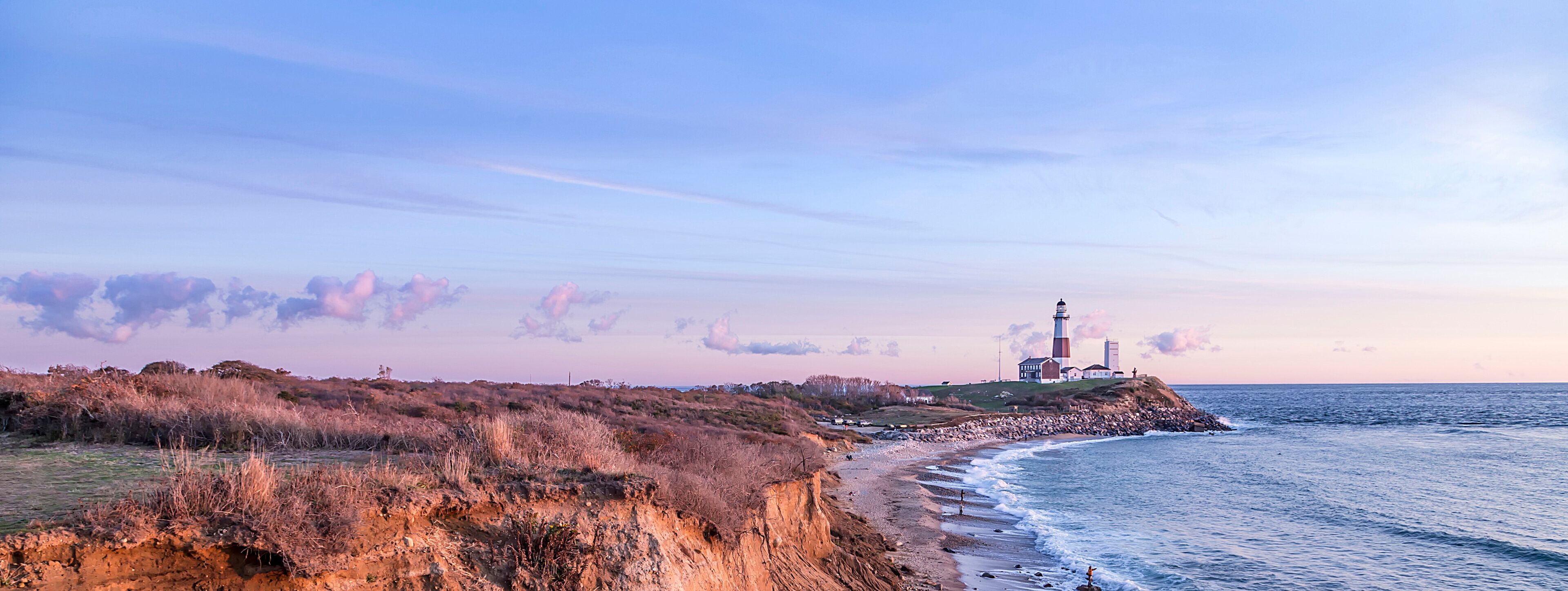 A coastal landscape with a lighthouse, grassy dunes, and a rocky shoreline.