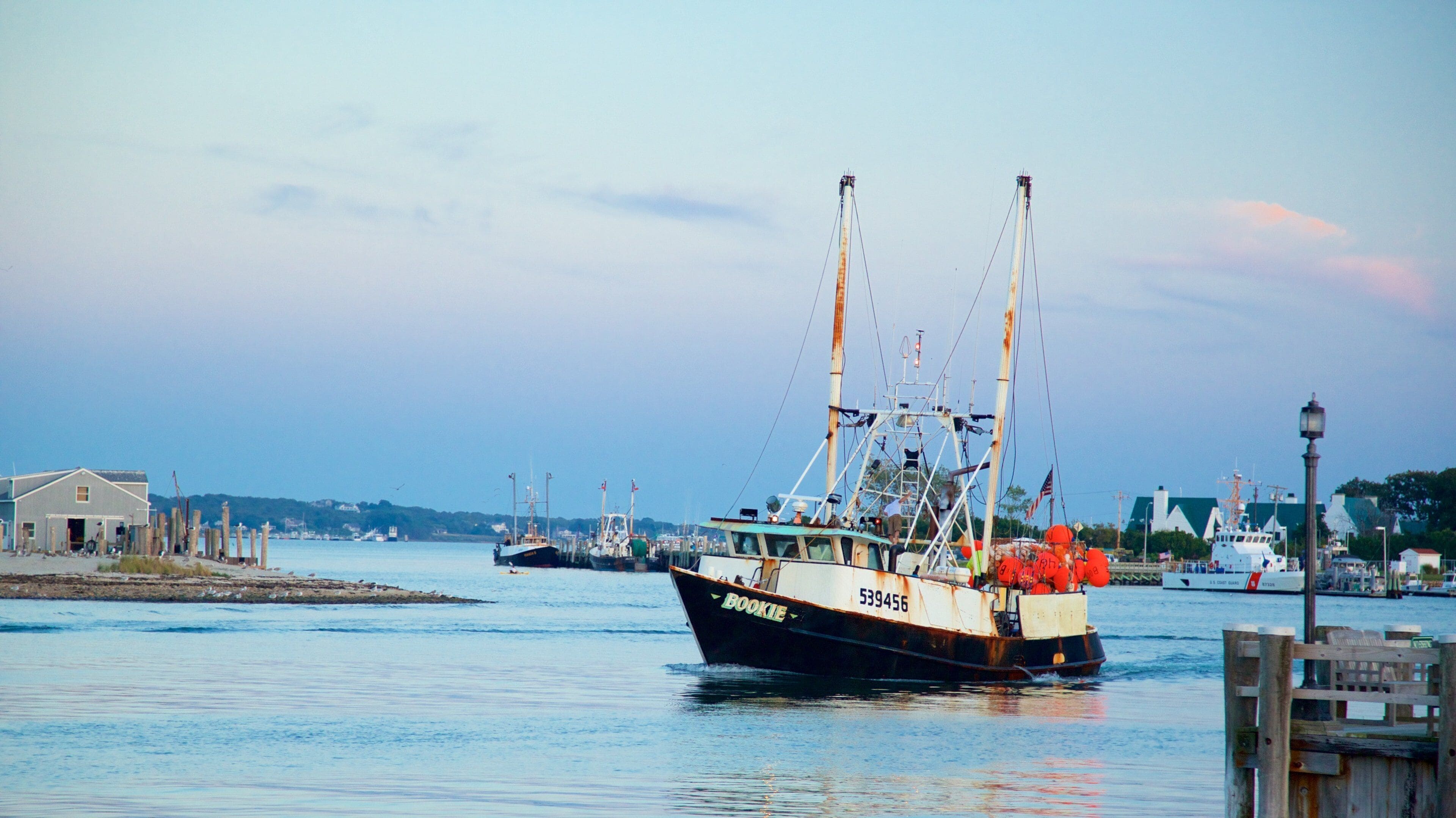 Un bateau de pêche nommé « Bookie », portant le numéro 539456, était amarré dans un port, à côté d’autres bateaux, avec un petit bâtiment visible au loin.
