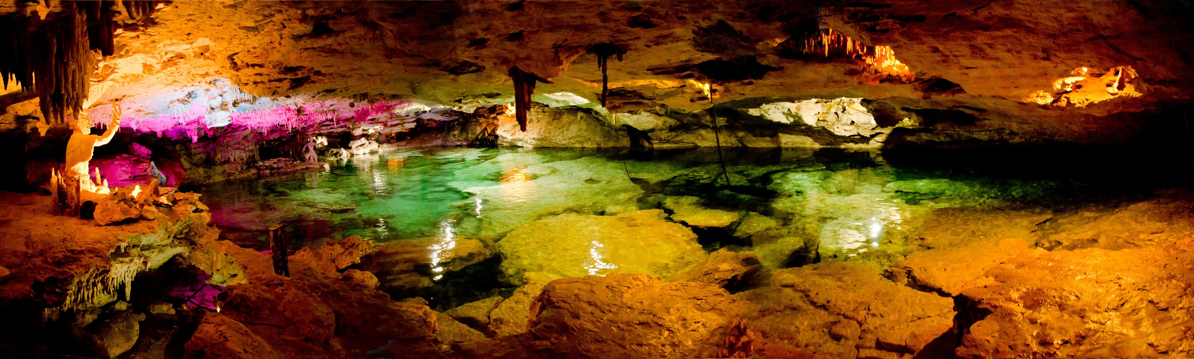 A cave with colorful stalactites and stalagmites, a pool of water, and a person exploring.