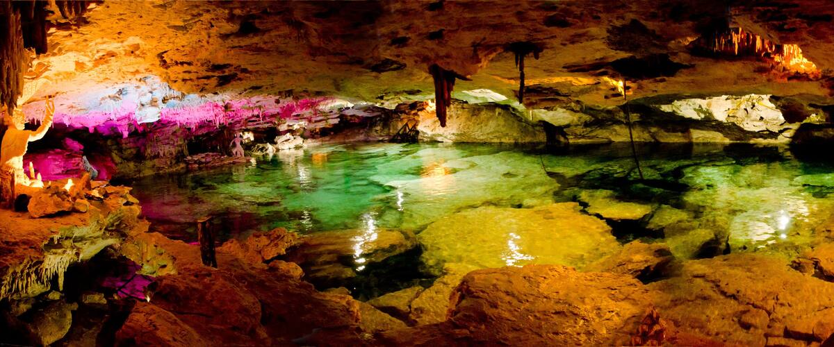 A cave with colorful stalactites and stalagmites, a pool of water, and a person exploring.